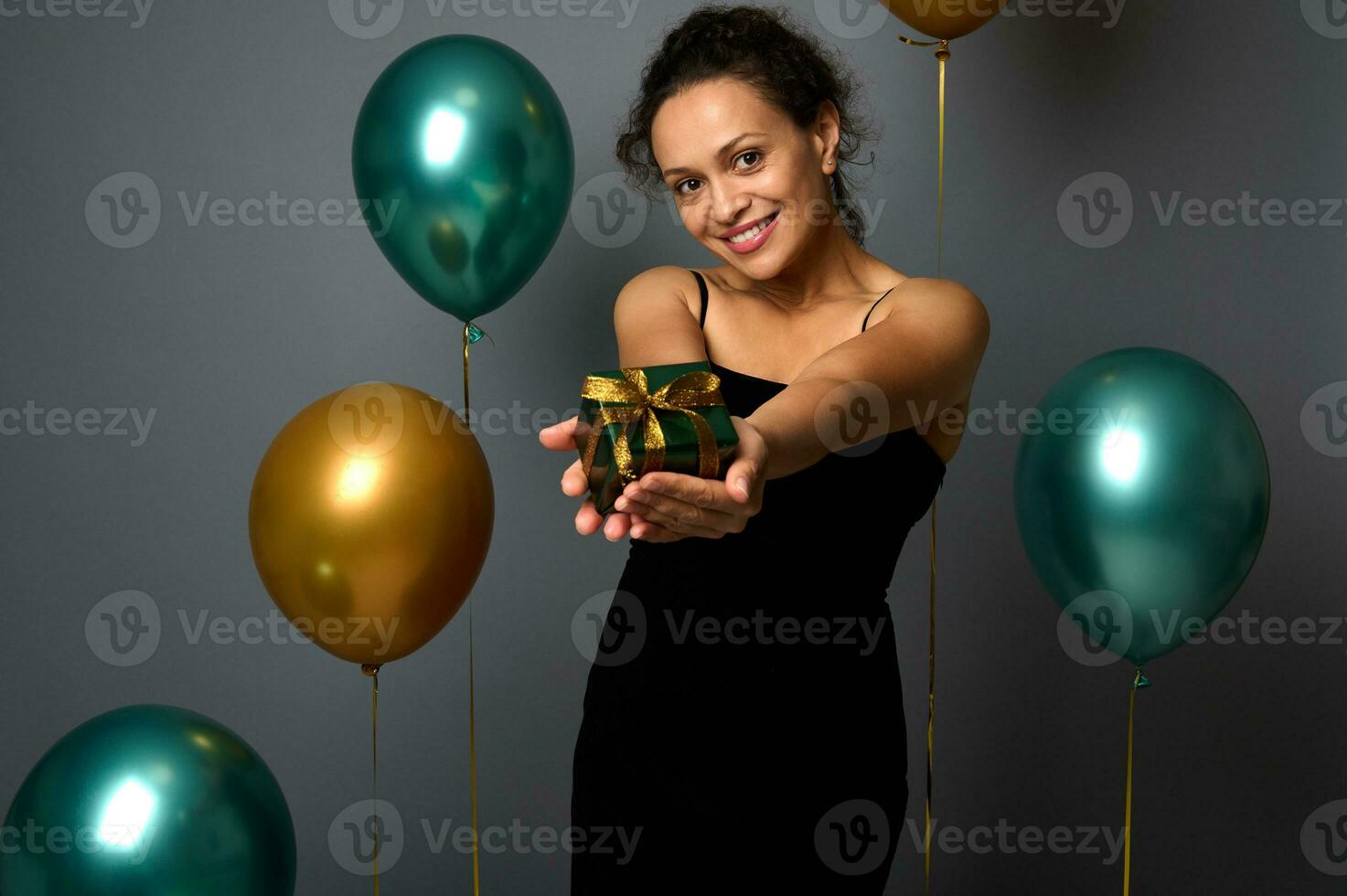 blandad lopp söt kvinna i svart klänning innehar en jul gåva i utsträckt händer och visar Det, ser på kamera, isolerat över grå bakgrund med guld och grön luft ballonger, kopia Plats för ad foto