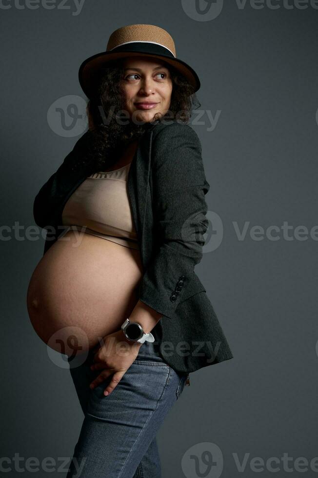 eleganta gravid kvinna med stor naken mage, klädd i jeans, blazer och sugrör hatt. de begrepp av skön graviditet foto