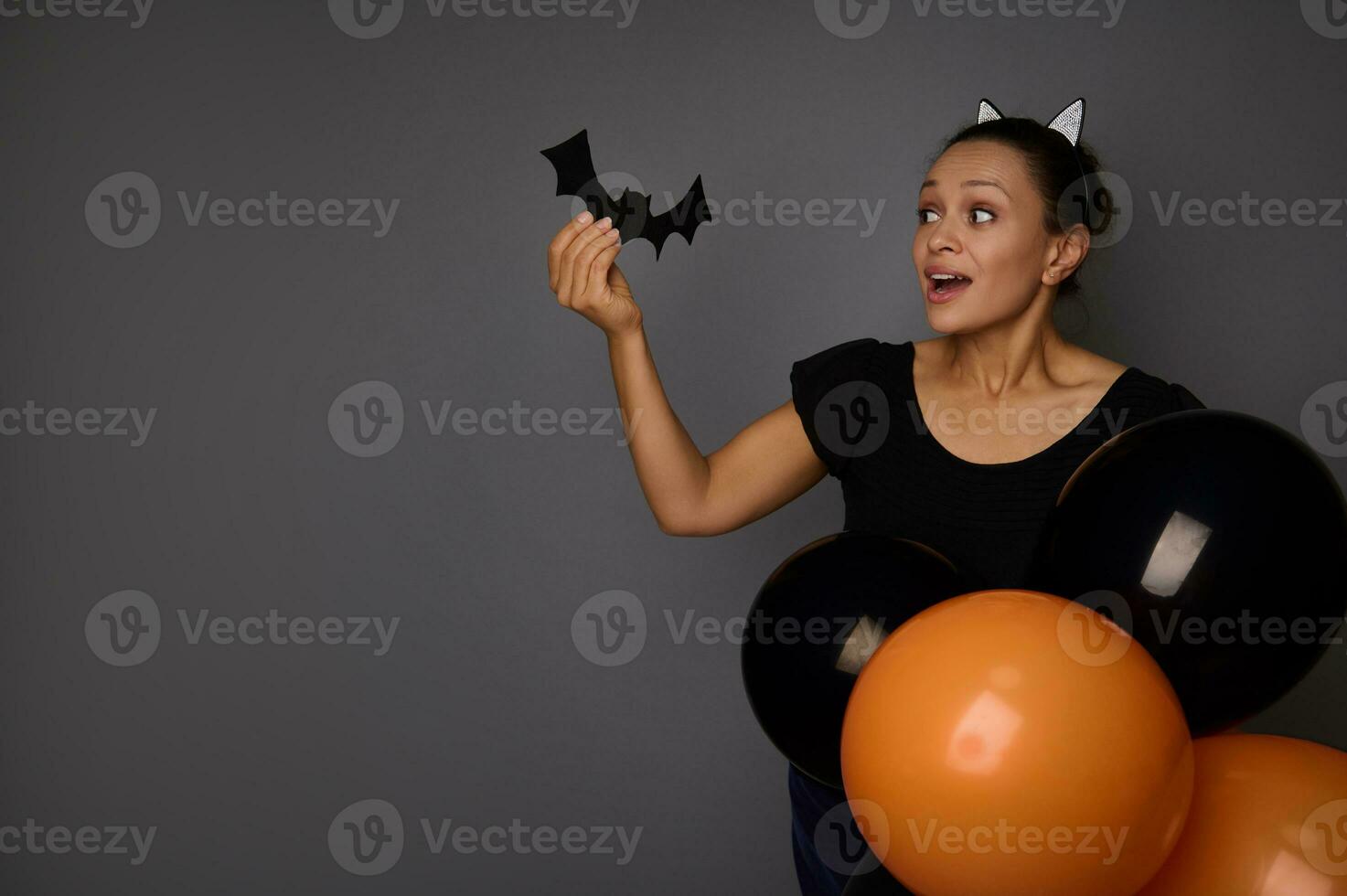 skön glad blandad lopp kvinna klädd i karneval kostym ser på en filtskuren handgjort fladdermus i henne hand , Framställ med färgrik svart orange luft ballonger mot grå bakgrund med kopia Plats foto