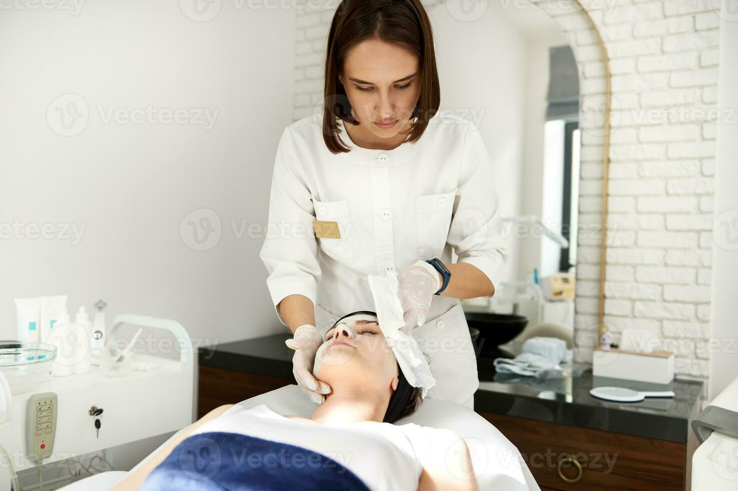 läkare kosmetolog utför skönhet procedur på kvinna ansikte, avlägsnande de resten av alginat mask använder sig av särskild våt servetter. professionell hud vård i kosmetologi klinik foto