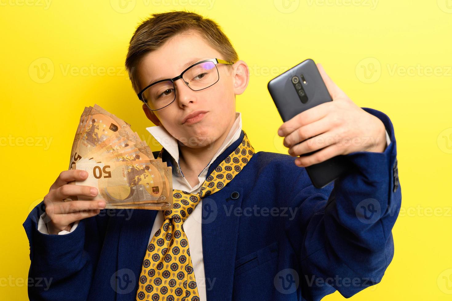 framgångsrikt och rikt barn som tar en selfie med pengar, porträtt av en pojke i kostym på gul bakgrund. foto