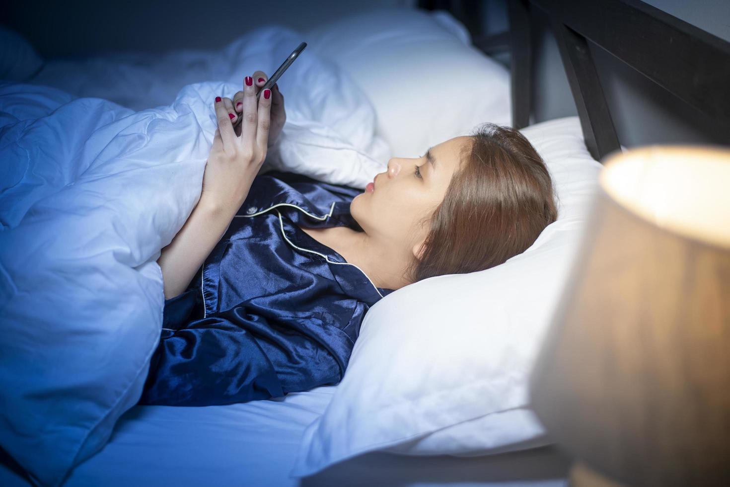 vacker kvinna spelar smart telefon innan du sover i sovrummet foto