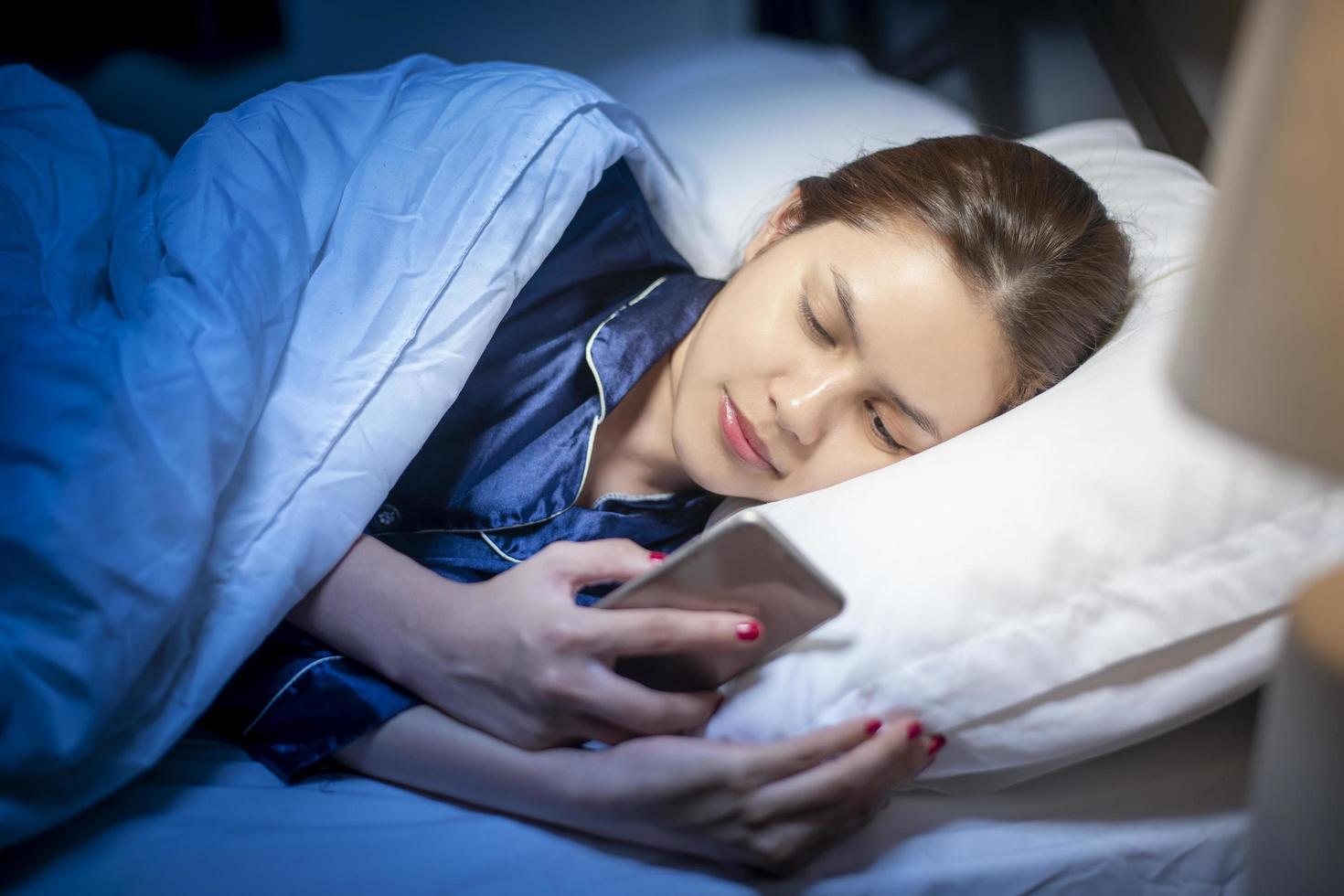 vacker kvinna spelar smart telefon innan du sover i sovrummet foto