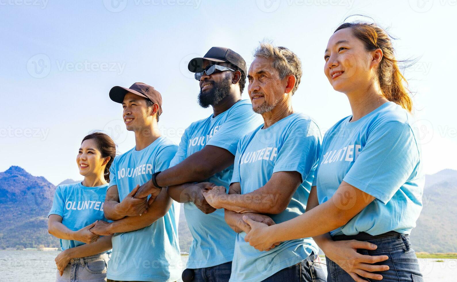 team av ung och mångfald volontär- arbetare grupp njut av välgörenhet social arbete utomhus- i strand rengöring projekt bär blå t-shirt medan sammanfogning hand i montera enhet foto