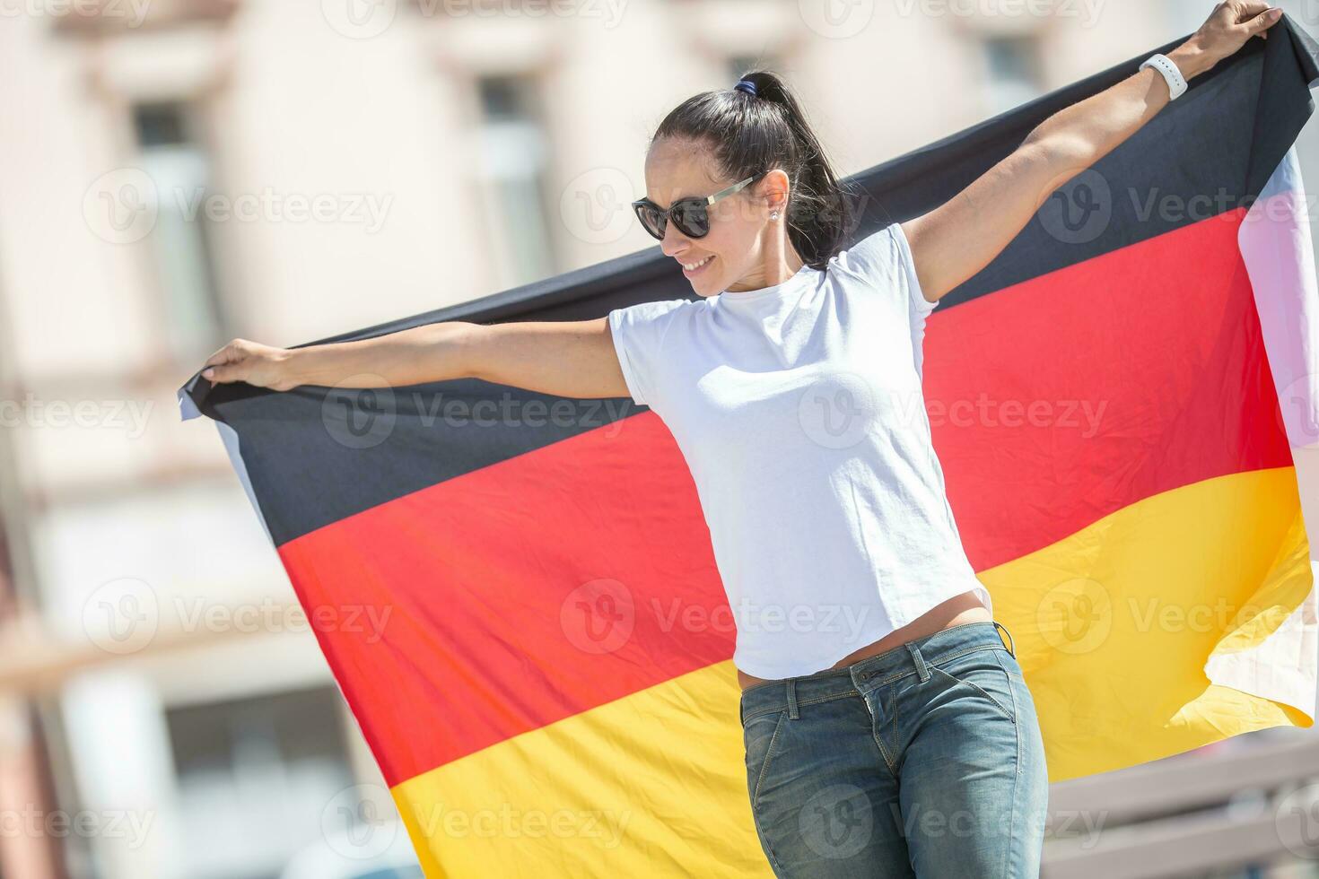 skön leende flicka i solglasögon innehar en tysk flagga Bakom henne utomhus foto