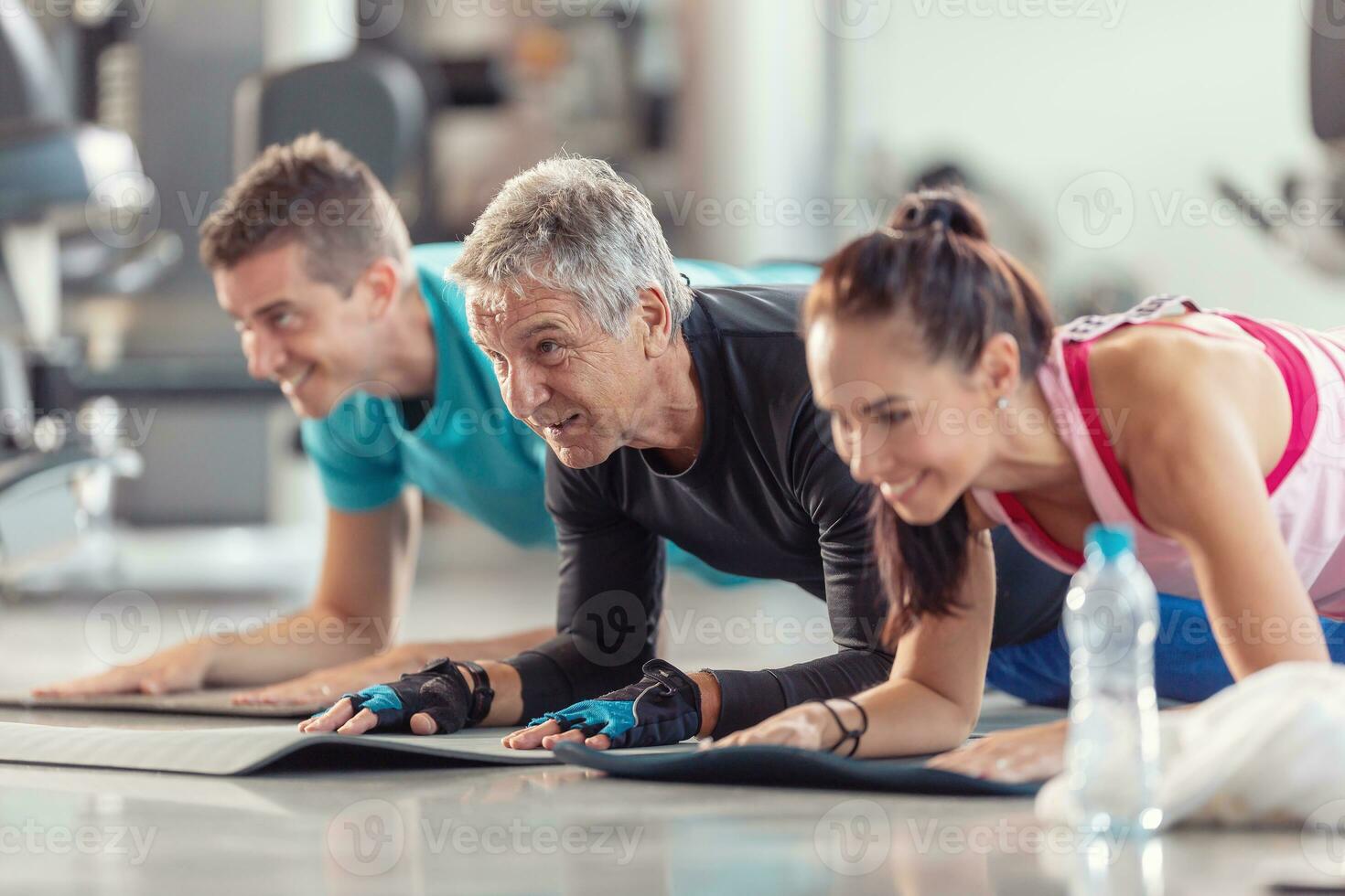 grupp av människor i olika ålder och kön har roligt medan håller på med grupp övning av armbåge planka i de Gym foto