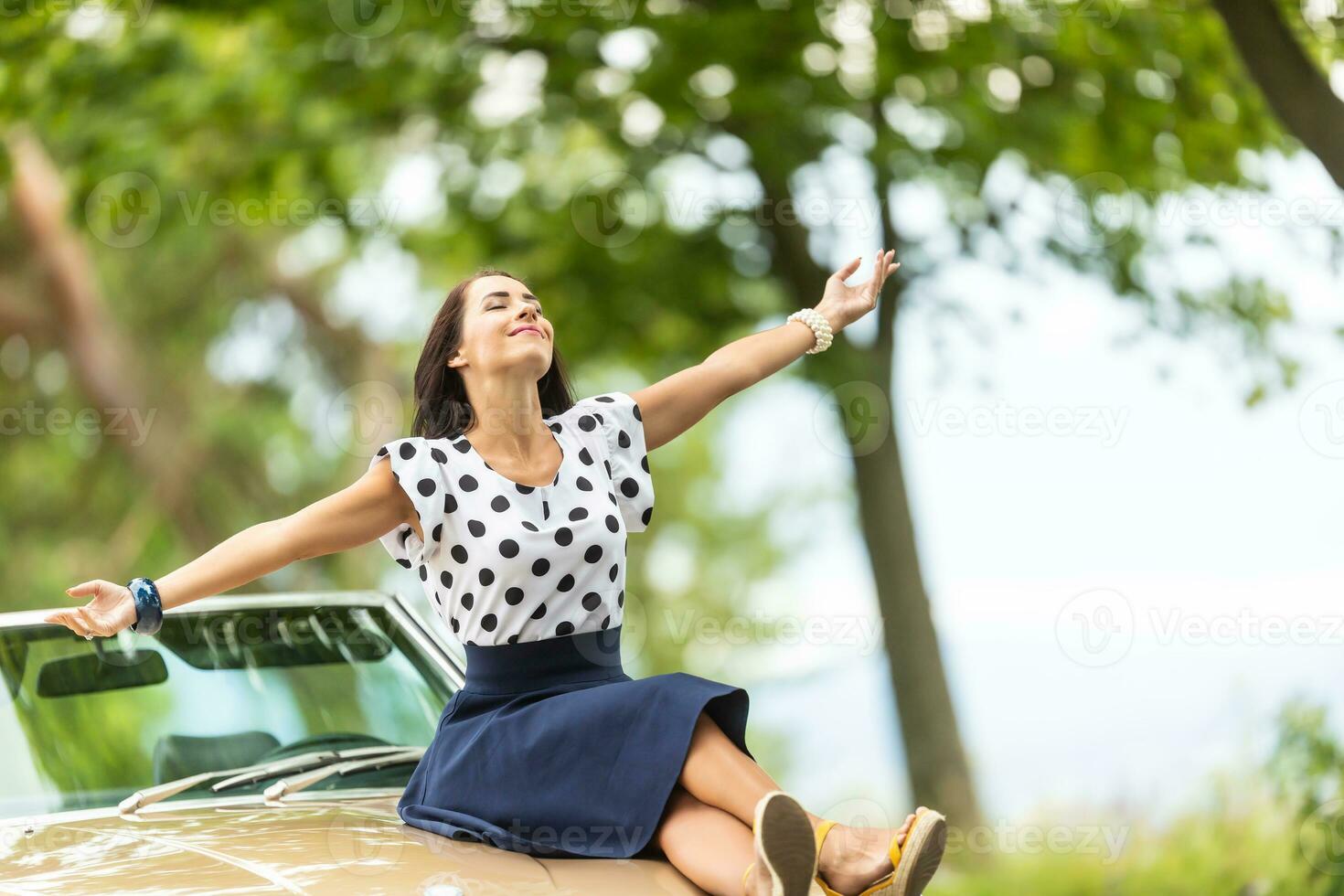 kvinna i polkadot blus och kjol sitter på en hätta av en cabrio bil, vapen öppna, njuter sommar foto