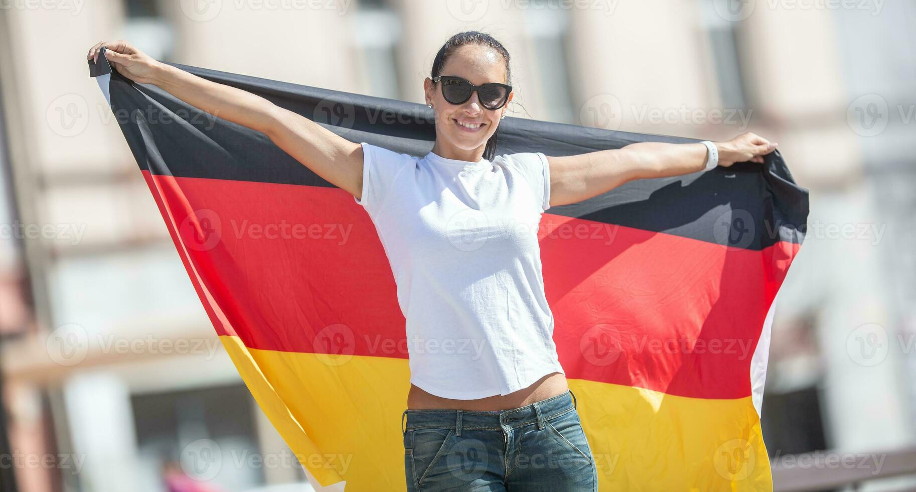 skön leende kvinna fläkt i solglasögon innehar en tysk flagga Bakom henne utomhus foto