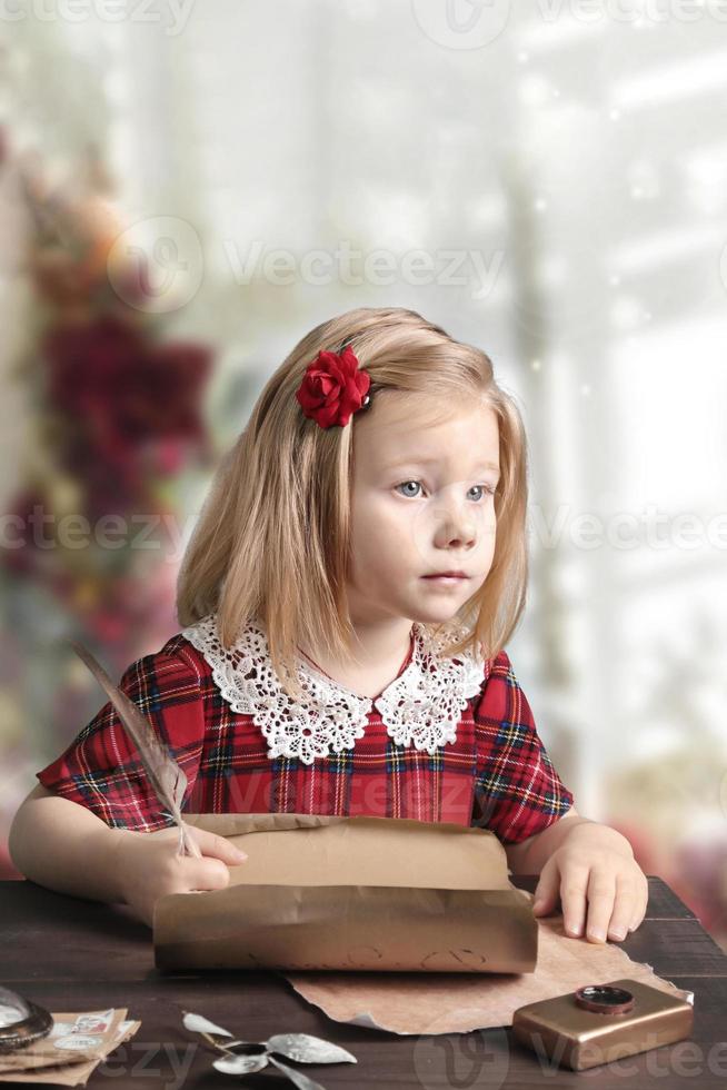 en liten flicka i en röd klänning sitter vid bordet och skriver ett brev till santa foto