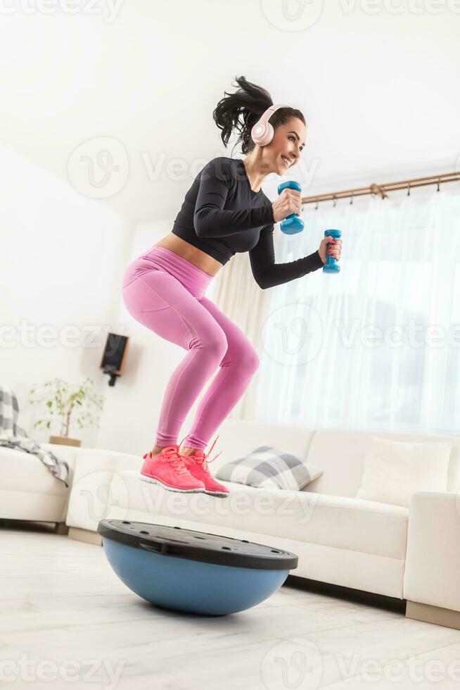 passa flicka i rosa yoga byxor och svart skjorta bär hörlurar och gör träna på Hem med blå hantlar och balans boll foto