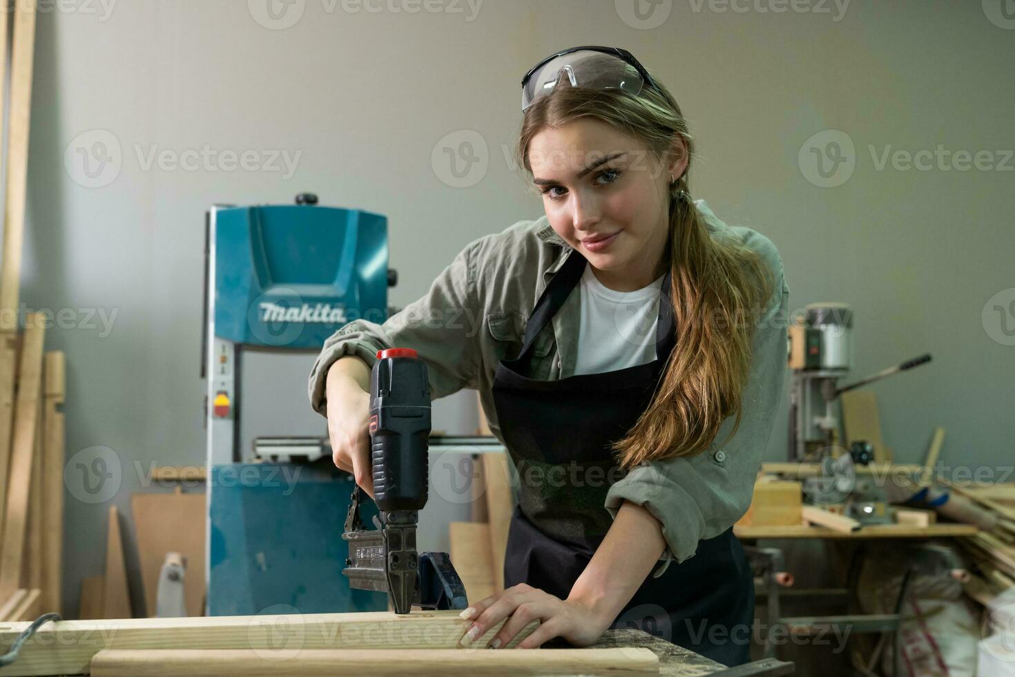 hårt arbetande snickare kvinna använder sig av verktyg leende självsäkert ung kvinna snickare i förkläde stående nära arbetsbänk och ser på kamera vänlig medan arbetssätt i de hantverk verkstad foto