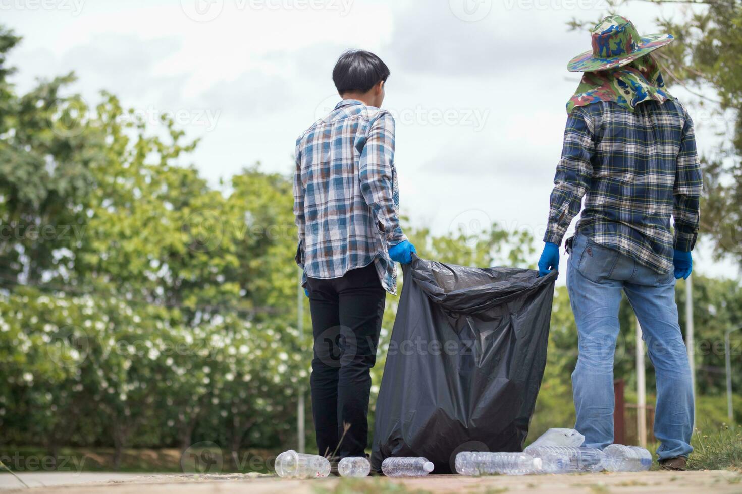 två man anställda använda sig av svart sopor påsar till samla plast flaskor och återvinningsbar avfall från de gräsmatta och trottoarer för återvinning. begrepp av sortering plast avfall för återvinning foto