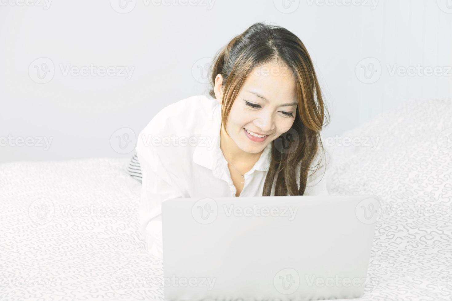 asiatisk kvinna som använder bärbar dator i emotionellt leende medan hon lägger på sängen. livsstil, avkoppling, fritid, teknik kommunikation koncept. foto