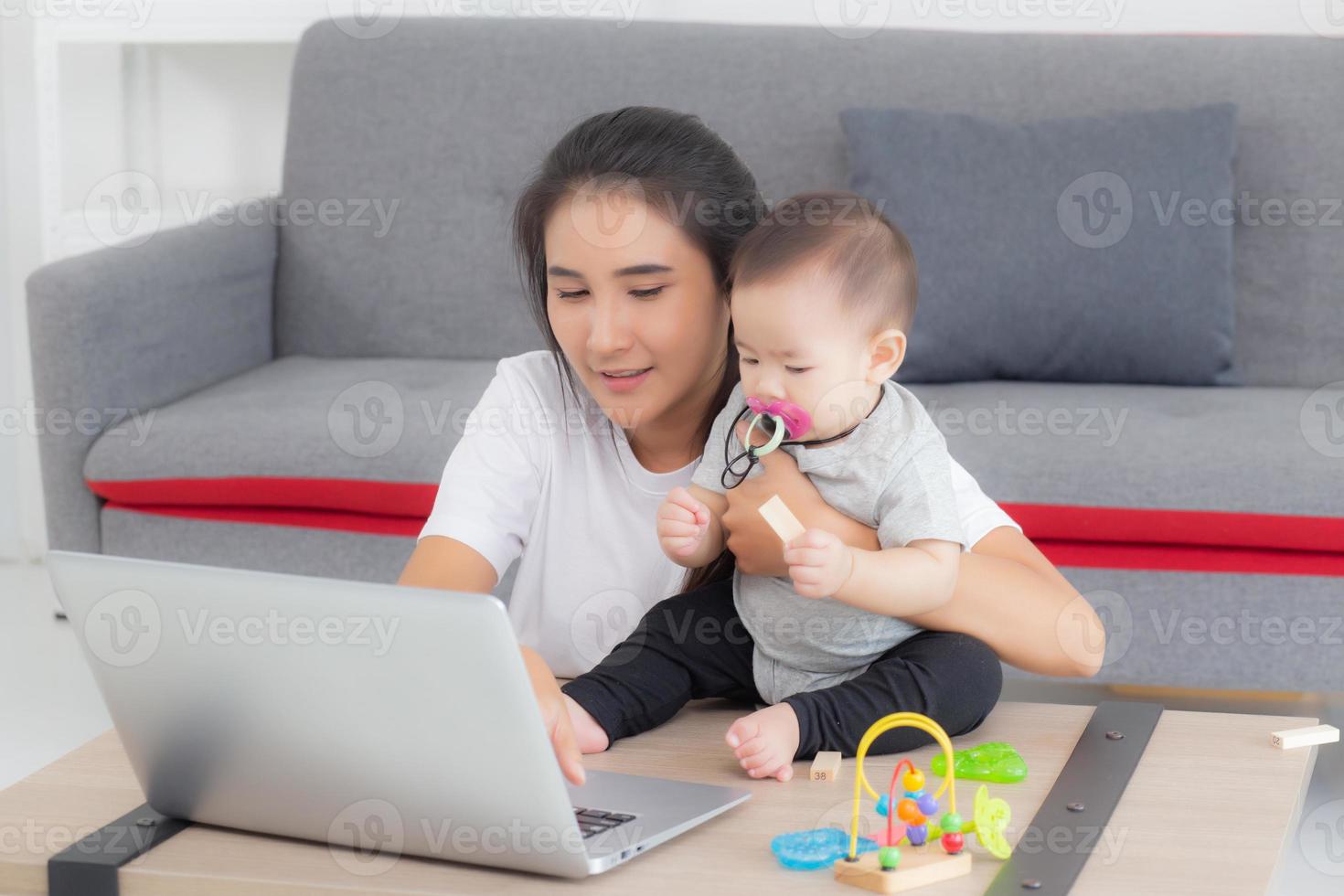 ung asiatisk mamma som arbetar på bärbar dator medan vårddotterflicka hemma, mamma och flickasammanträde med anteckningsbokvideosamtal, förälder och limning, inomhus, familj och affärsidé. foto