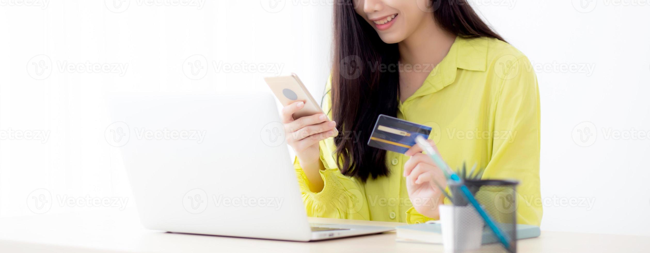 ung asiatisk affärskvinna som använder den smarta telefonen och innehar kreditkortet medan online-shopping och betalning online med bärbar dator på skrivbordet hemma, kvinnligt innehavbetalkort, kommunikationskoncept. foto