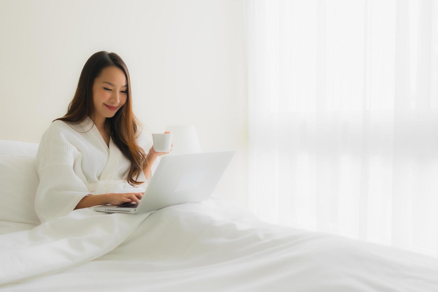 stående vackra unga asiatiska kvinnor med kaffekopp och datorbärbar dator på sängen foto
