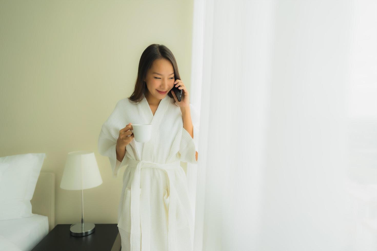 stående vackra unga asiatiska kvinnor med kaffekoppen och mobiltelefonen på sängen foto