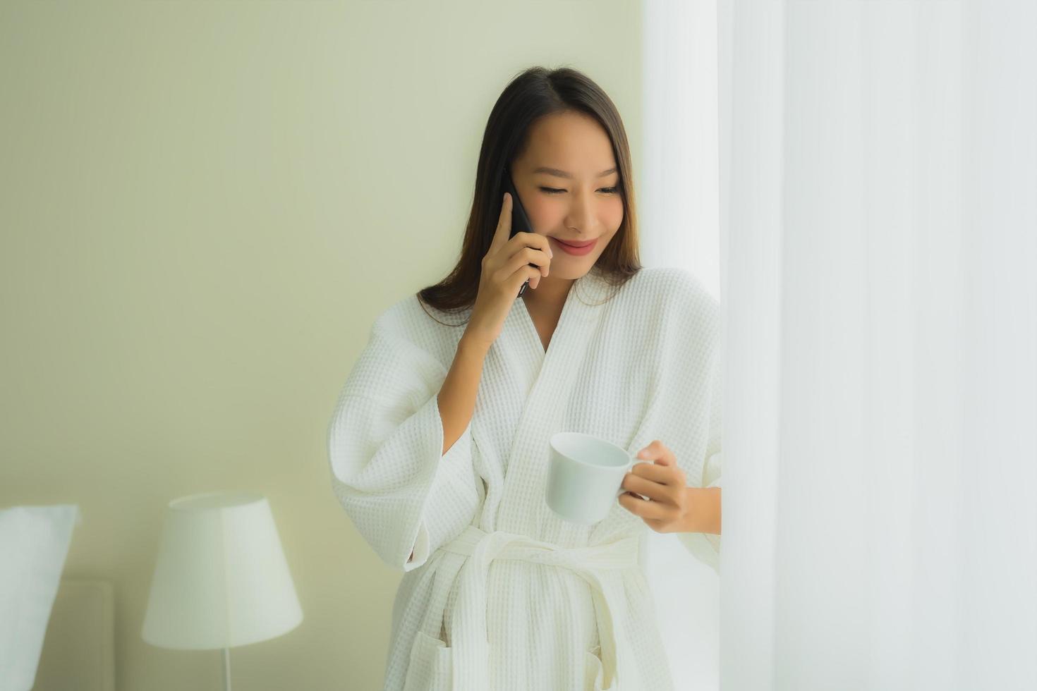 stående vackra unga asiatiska kvinnor med kaffekoppen och mobiltelefonen på sängen foto