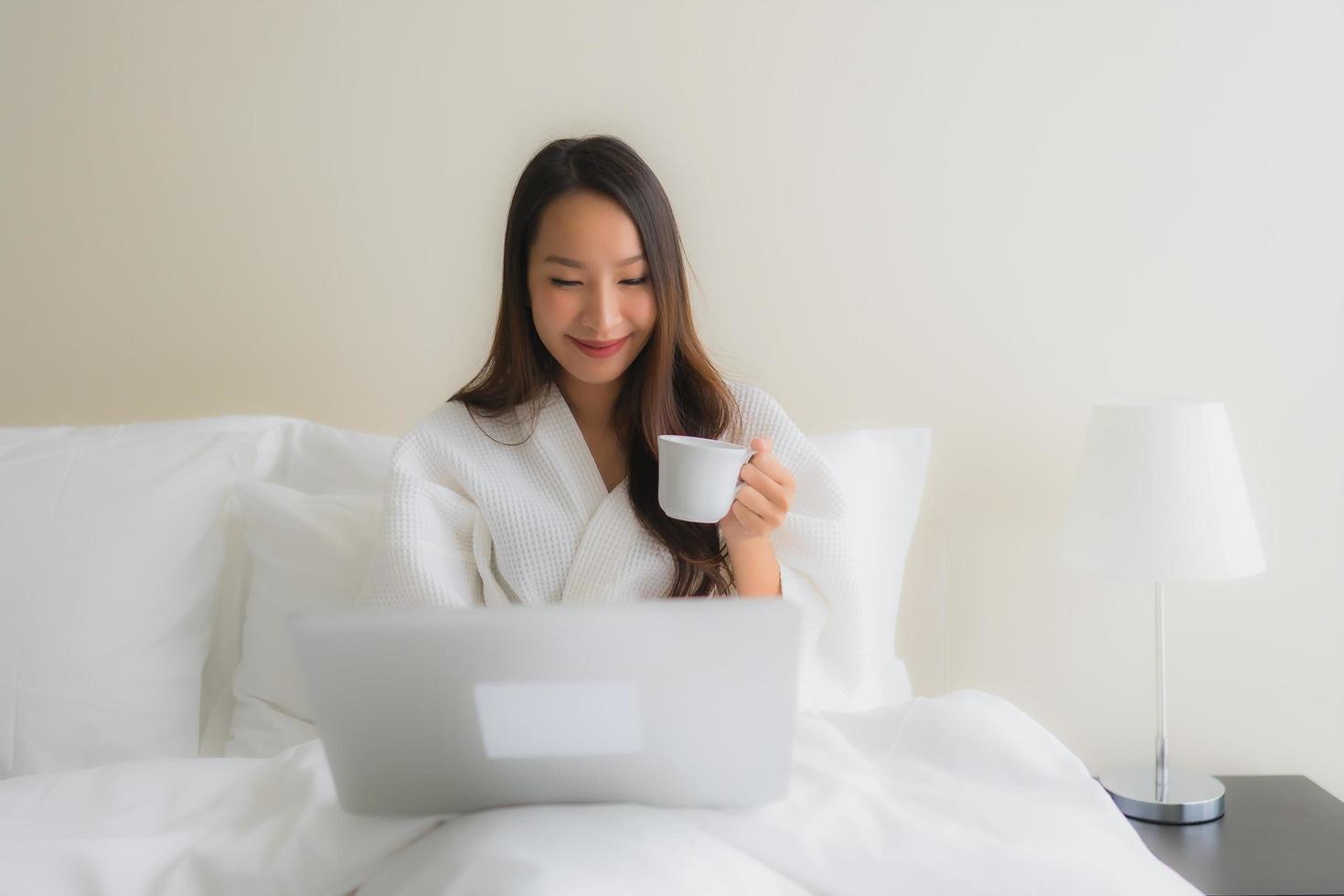 stående vackra unga asiatiska kvinnor med kaffekopp och datorbärbar dator på sängen foto