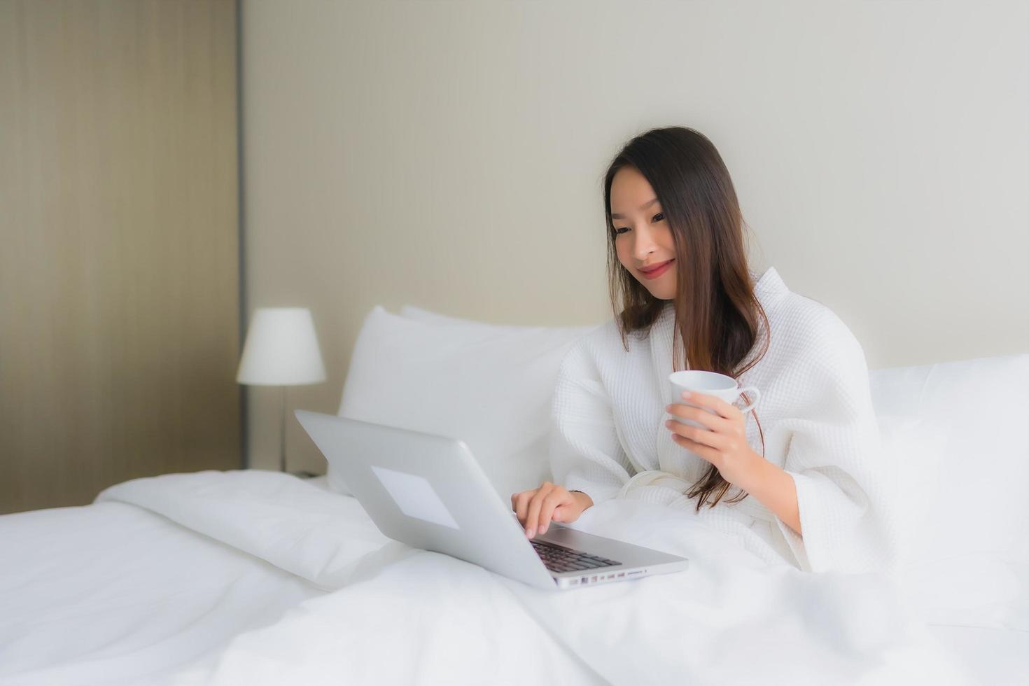 stående vackra unga asiatiska kvinnor med kaffekopp och datorbärbar dator på sängen foto
