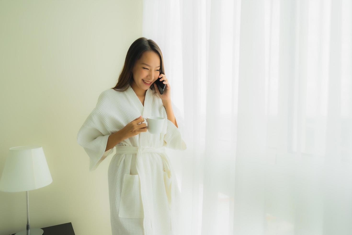 stående vackra unga asiatiska kvinnor med kaffekoppen och mobiltelefonen på sängen foto