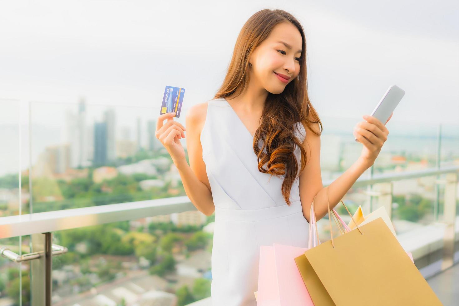 stående vacker ung asiatisk kvinna glad och ler med kreditkort och mobil eller smart telefon och shoppingväska foto