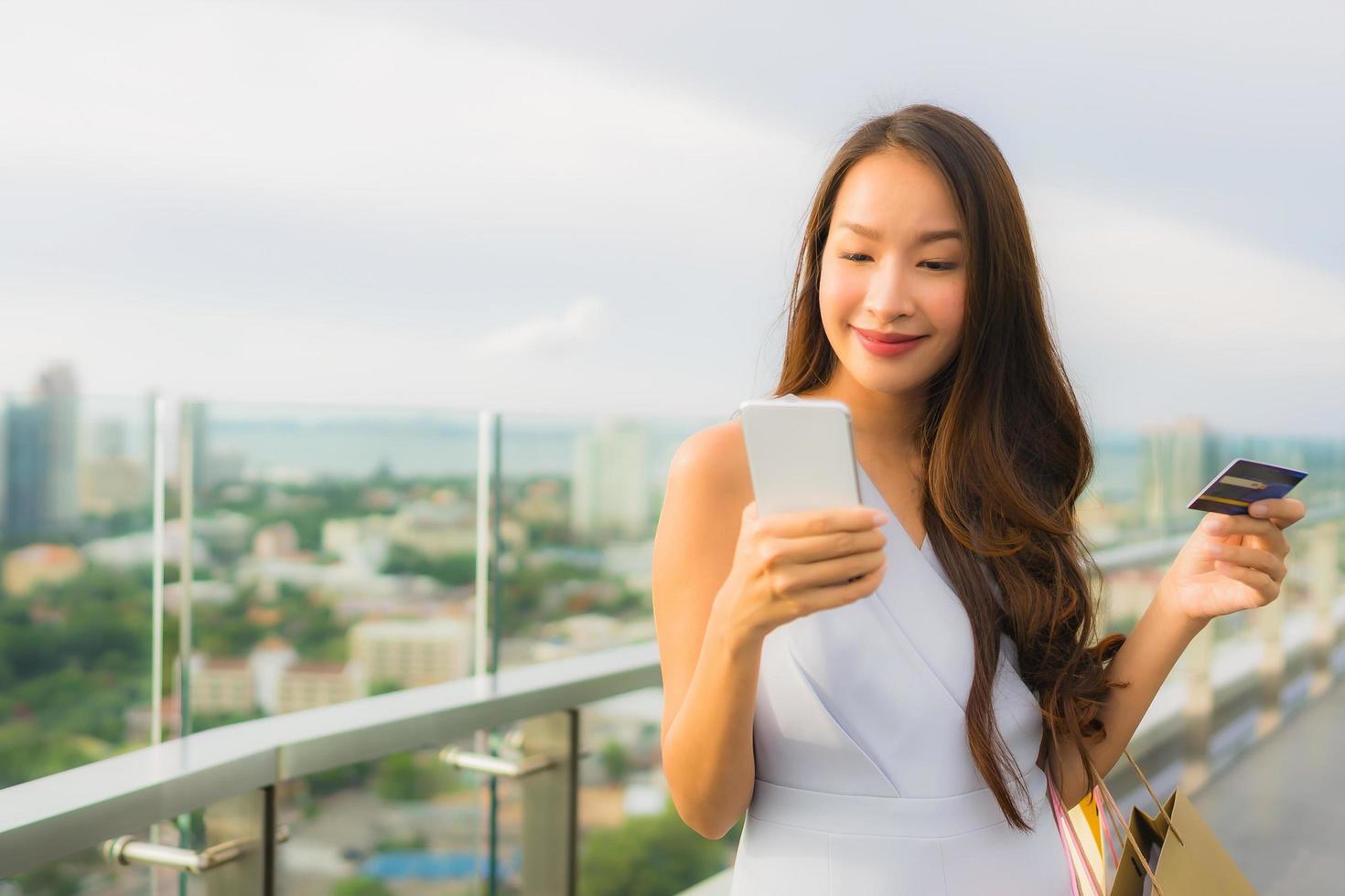 stående vacker ung asiatisk kvinna glad och ler med kreditkort och mobil eller smart telefon och shoppingväska foto