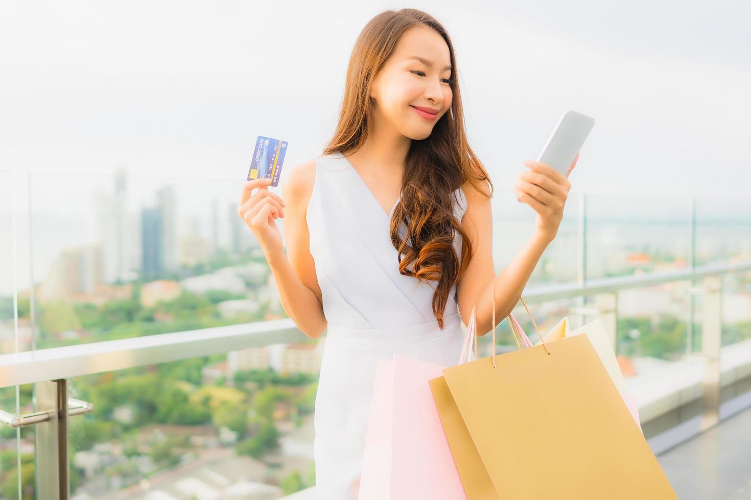 stående vacker ung asiatisk kvinna glad och ler med kreditkort och mobil eller smart telefon och shoppingväska foto