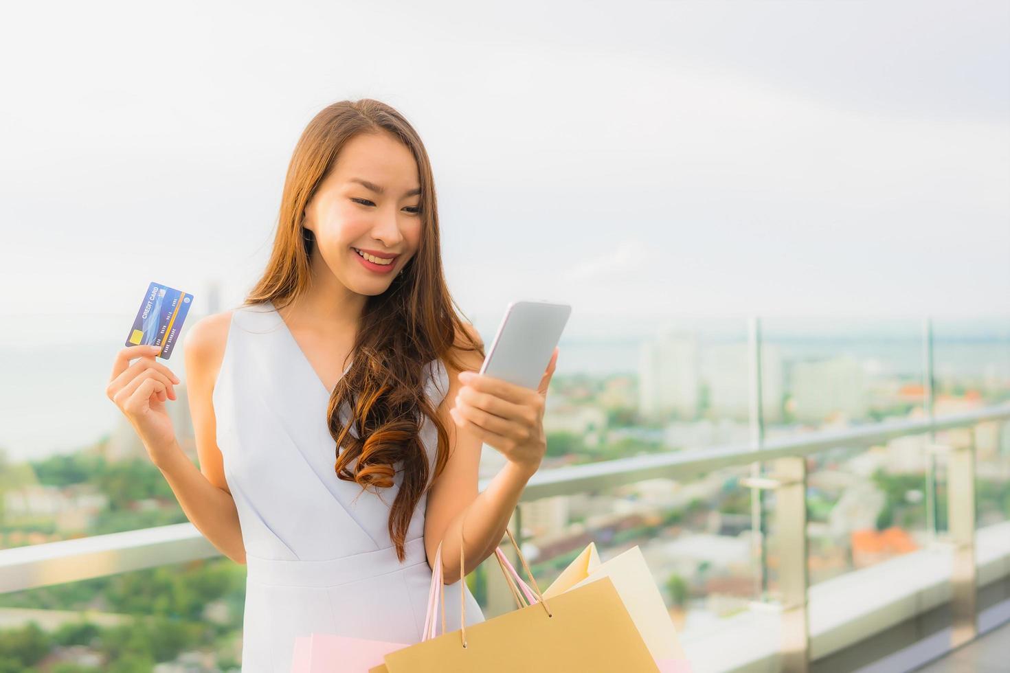 stående vacker ung asiatisk kvinna glad och ler med kreditkort och mobil eller smart telefon och shoppingväska foto