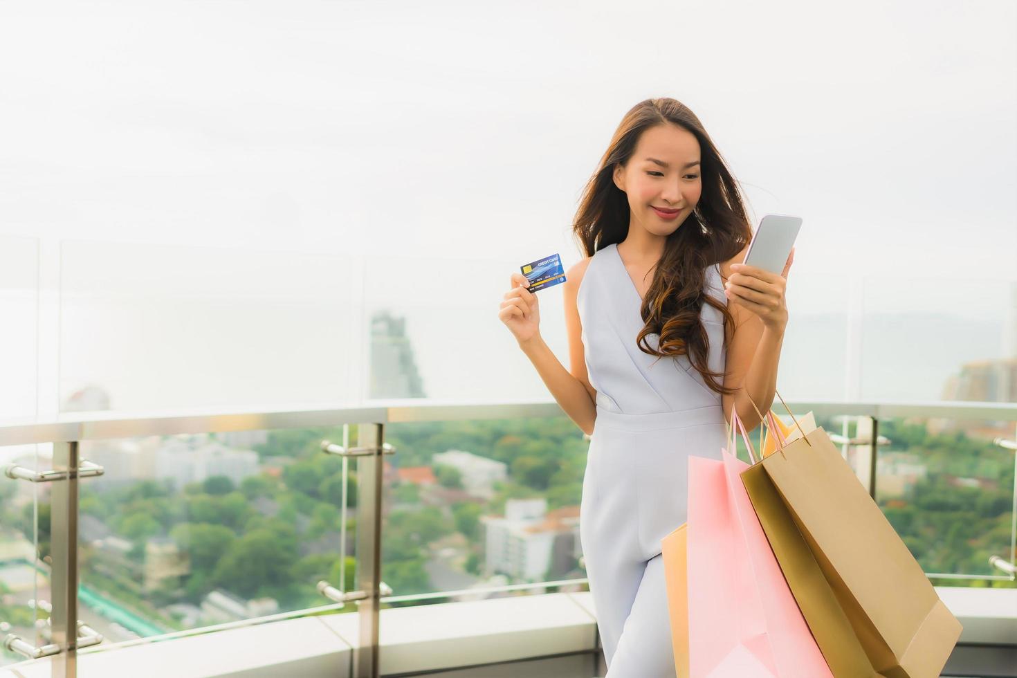 stående vacker ung asiatisk kvinna glad och ler med kreditkort och mobil eller smart telefon och shoppingväska foto