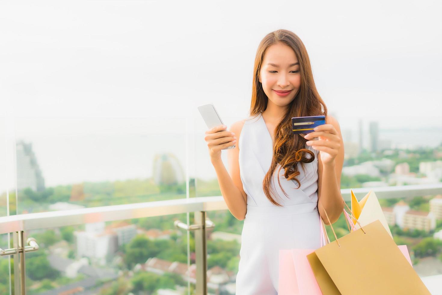 stående vacker ung asiatisk kvinna glad och ler med kreditkort och mobil eller smart telefon och shoppingväska foto