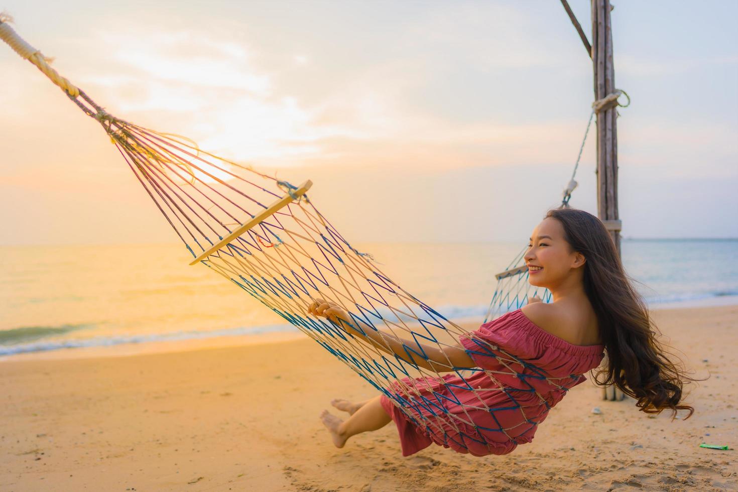stående vacker ung asiatisk kvinna som sitter på hängmattan med leende glad neary strand hav och hav foto