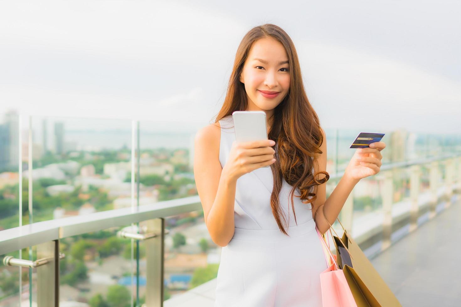 stående vacker ung asiatisk kvinna glad och ler med kreditkort och mobil eller smart telefon och shoppingväska foto