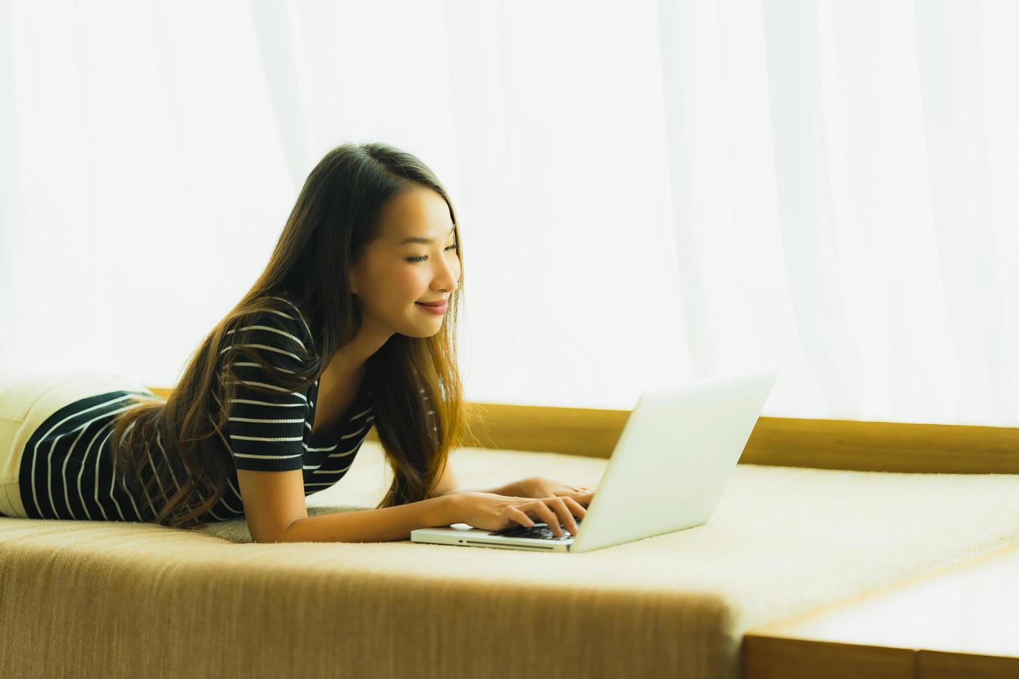 stående härlig ung asiatisk kvinna som använder datoranteckningsboken eller bärbar dator på soffan i vardagsrummet foto