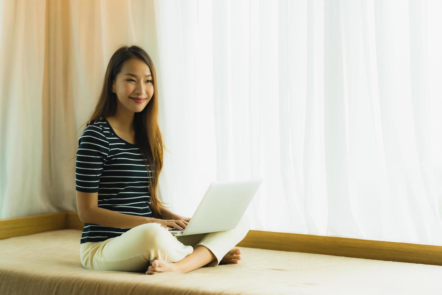 stående härlig ung asiatisk kvinna som använder datoranteckningsboken eller bärbar dator på soffan i vardagsrummet foto