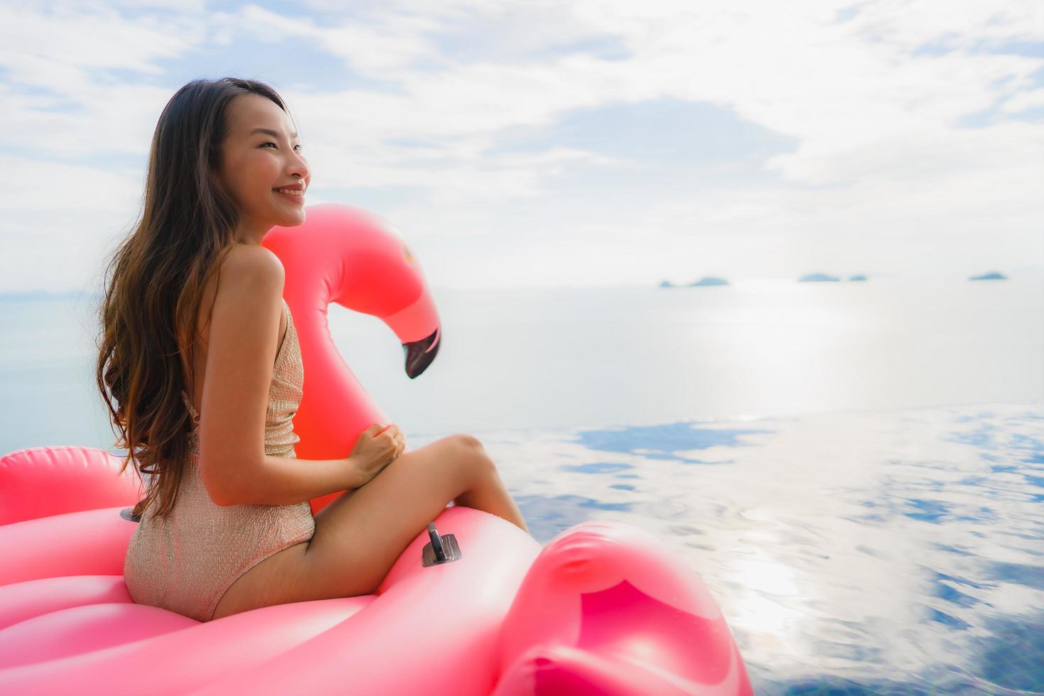 stående ung asiatisk kvinna på uppblåsbar flottörflamingo runt utomhuspool i hotellresort foto