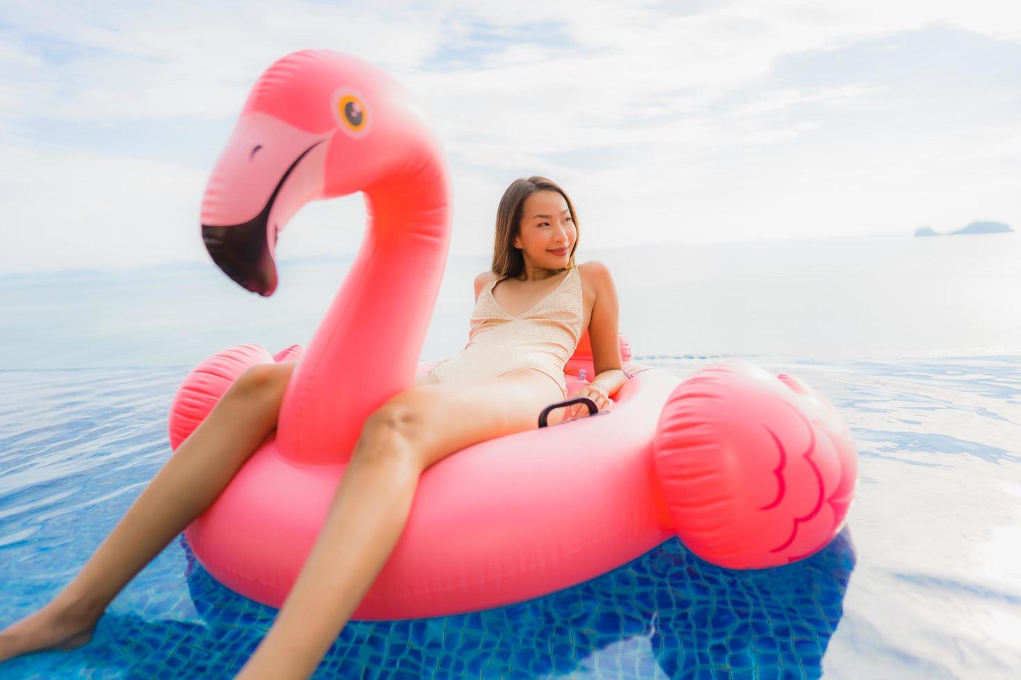 stående ung asiatisk kvinna på uppblåsbar flottörflamingo runt utomhuspool i hotellresort foto