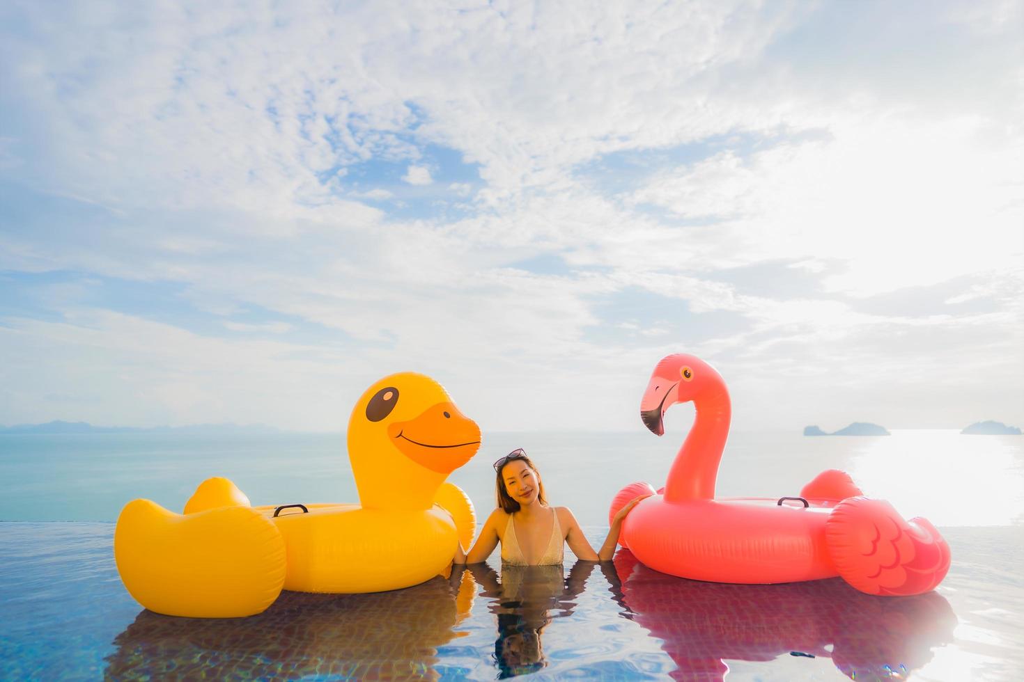 stående ung asiatisk kvinna på uppblåsbar flottör gul anka och rosa flamingo runt utomhuspool i hotell och semesterort foto