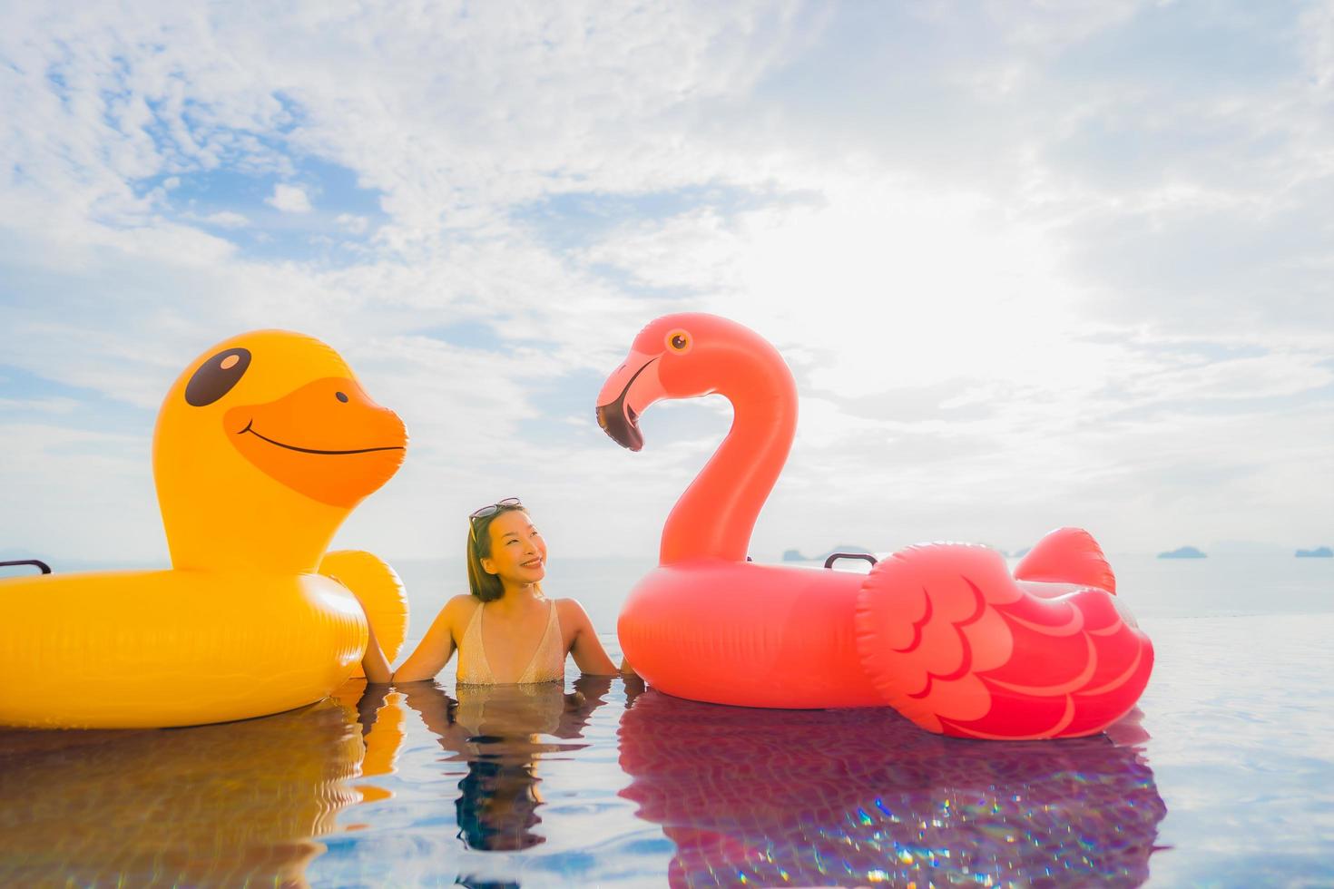 stående ung asiatisk kvinna på uppblåsbar flottör gul anka och rosa flamingo runt utomhuspool i hotell och semesterort foto