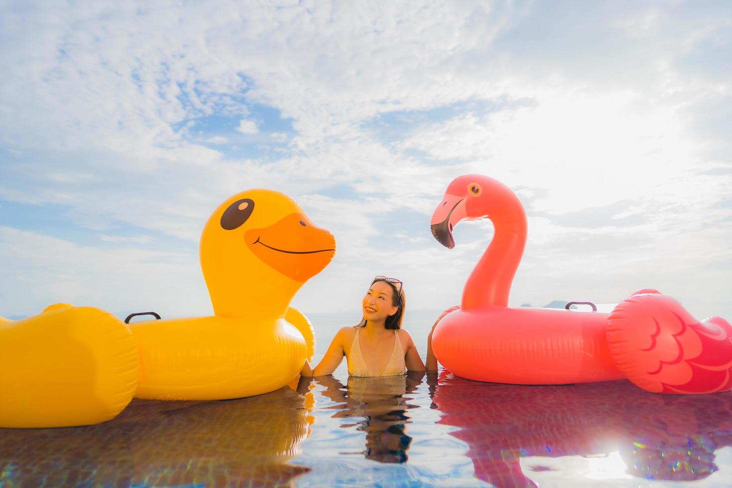 stående ung asiatisk kvinna på uppblåsbar flottör gul anka och rosa flamingo runt utomhuspool i hotell och semesterort foto