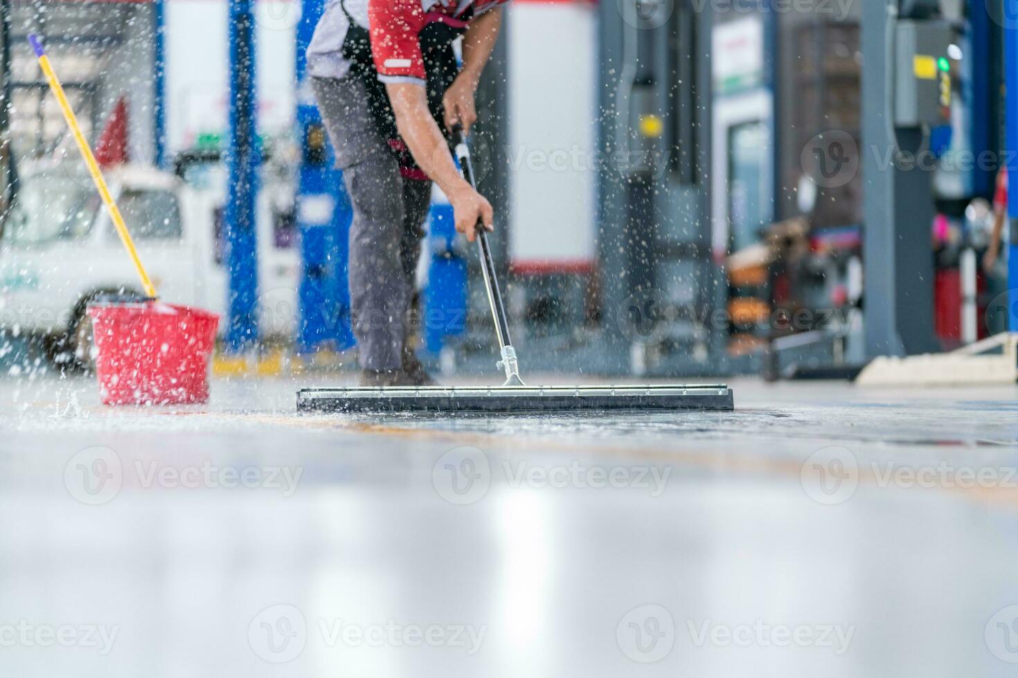 service personal man använder sig av en mopp till ta bort vatten i de enhetlig rengöring de skyddande Kläder av de ny epoxi golv i ett tömma lager eller bil service Centrum. foto