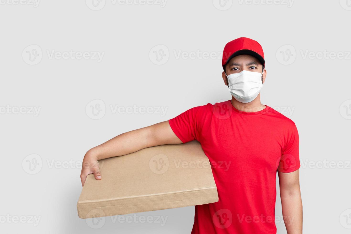 leverans man anställd i röd mössa tom t-shirt enhetlig ansiktsmask hålla Tom kartong isolerad på vit bakgrund foto