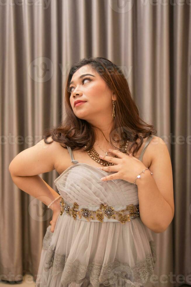 ett asiatisk kvinna bär en grå klänning och skön smink medan stående i främre av lyxig gardiner på en fest foto