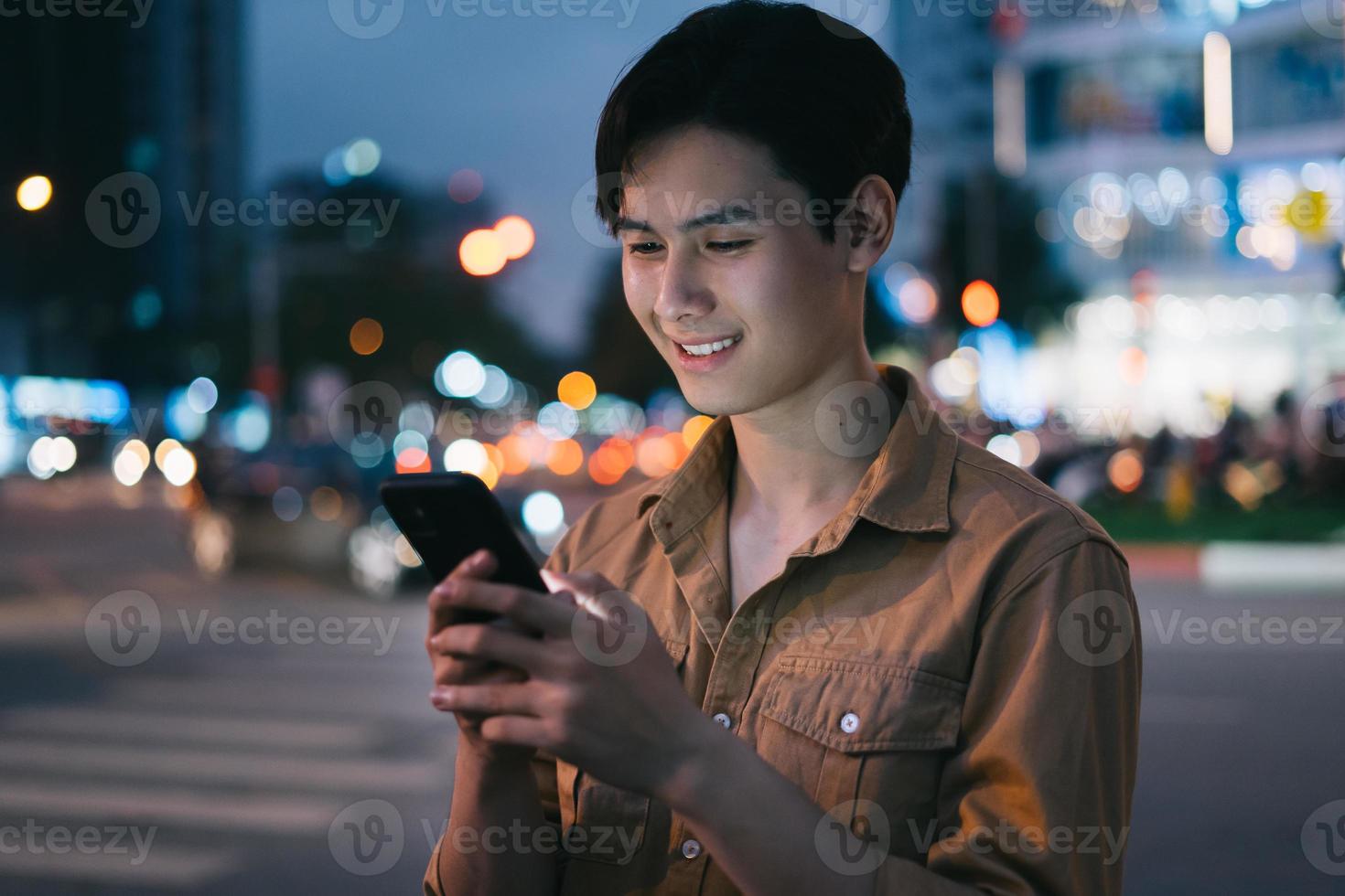 ung asiatisk man använder sin telefon medan han går på gatan på natten foto