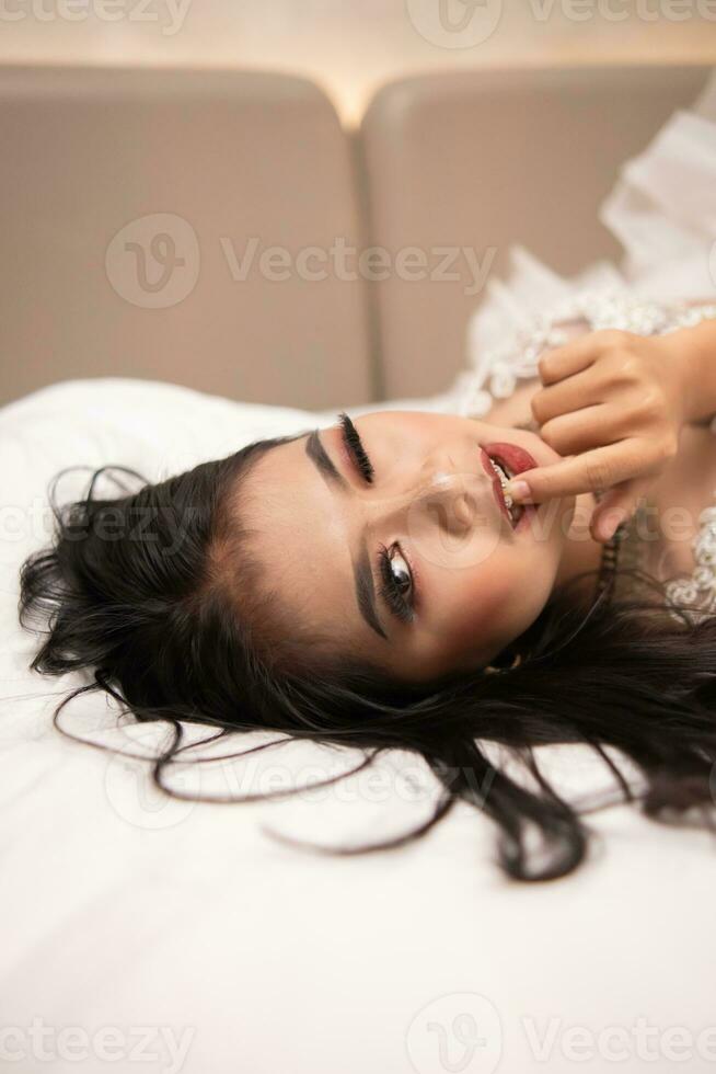 ett asiatisk kvinna med svart hår bitande henne fingrar med en kåt uttryck medan sovande på en vit säng foto