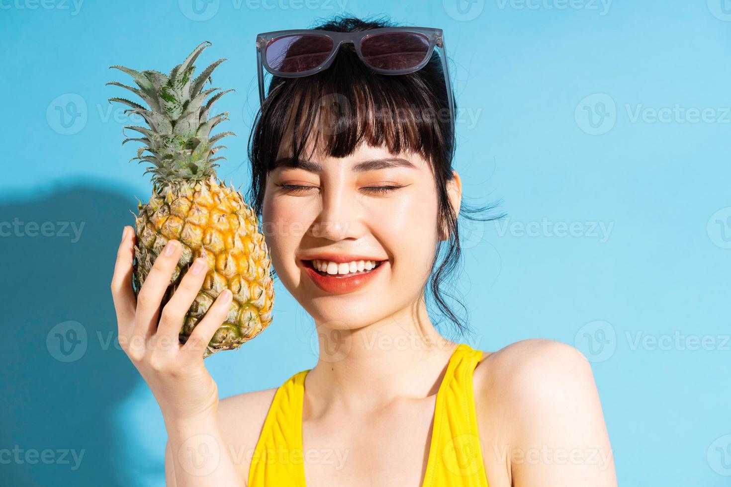 vacker asiatisk kvinna som bär gul jumpsuit på blå bakgrund och äter tropiska frukter, sommarbegrepp foto