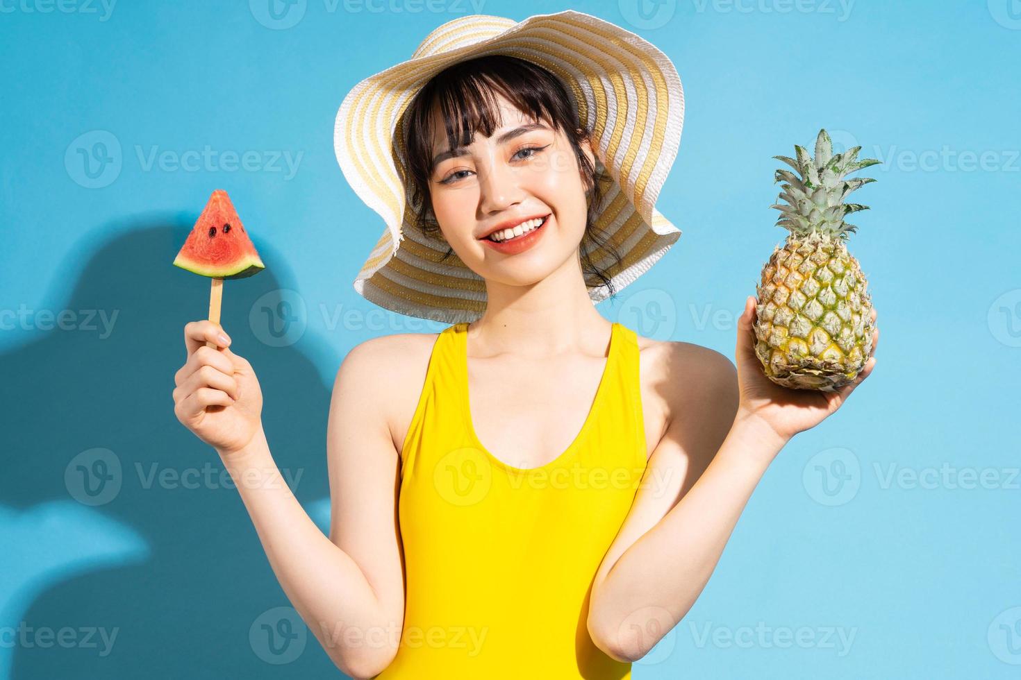 vacker asiatisk kvinna som bär gul jumpsuit på blå bakgrund och äter tropiska frukter, sommarbegrepp foto