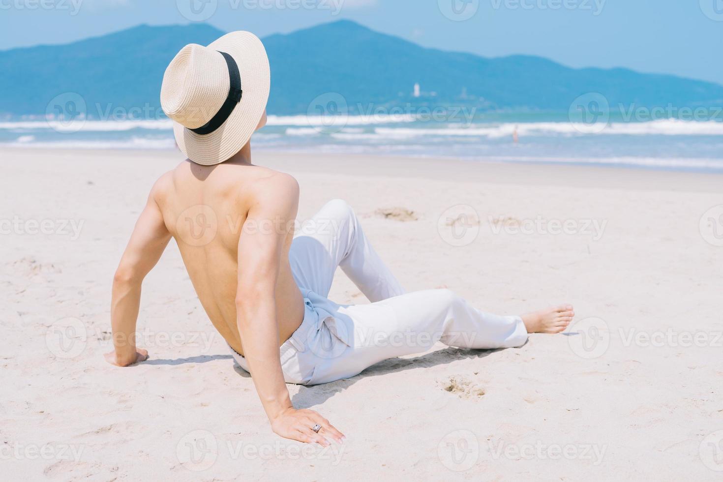 barbackad ung asiatisk man som sitter på sanden och tittar på havet foto