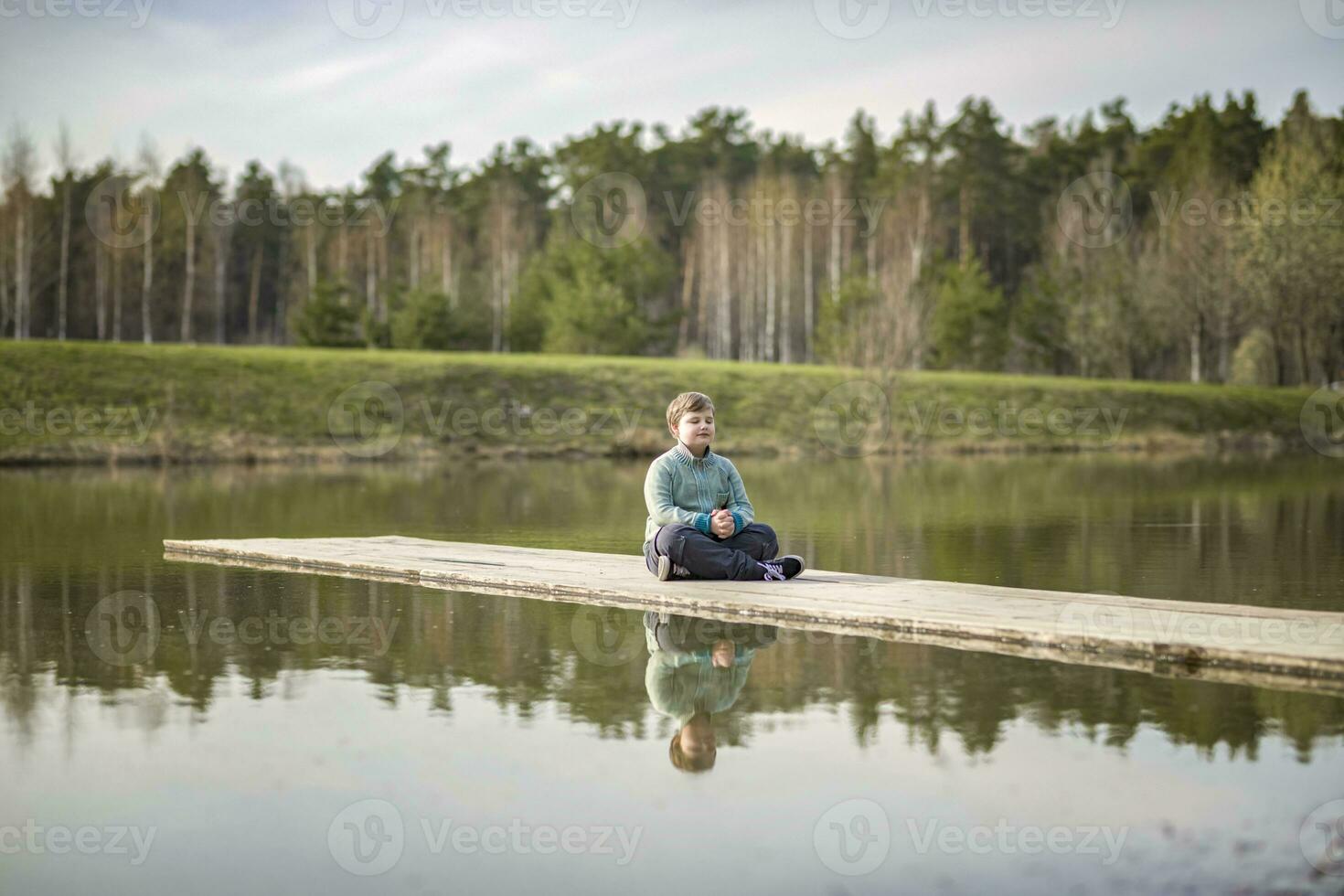 en pojke är Sammanträde på en bro i en grön parkera. de väg är en bro över de sjö. foto