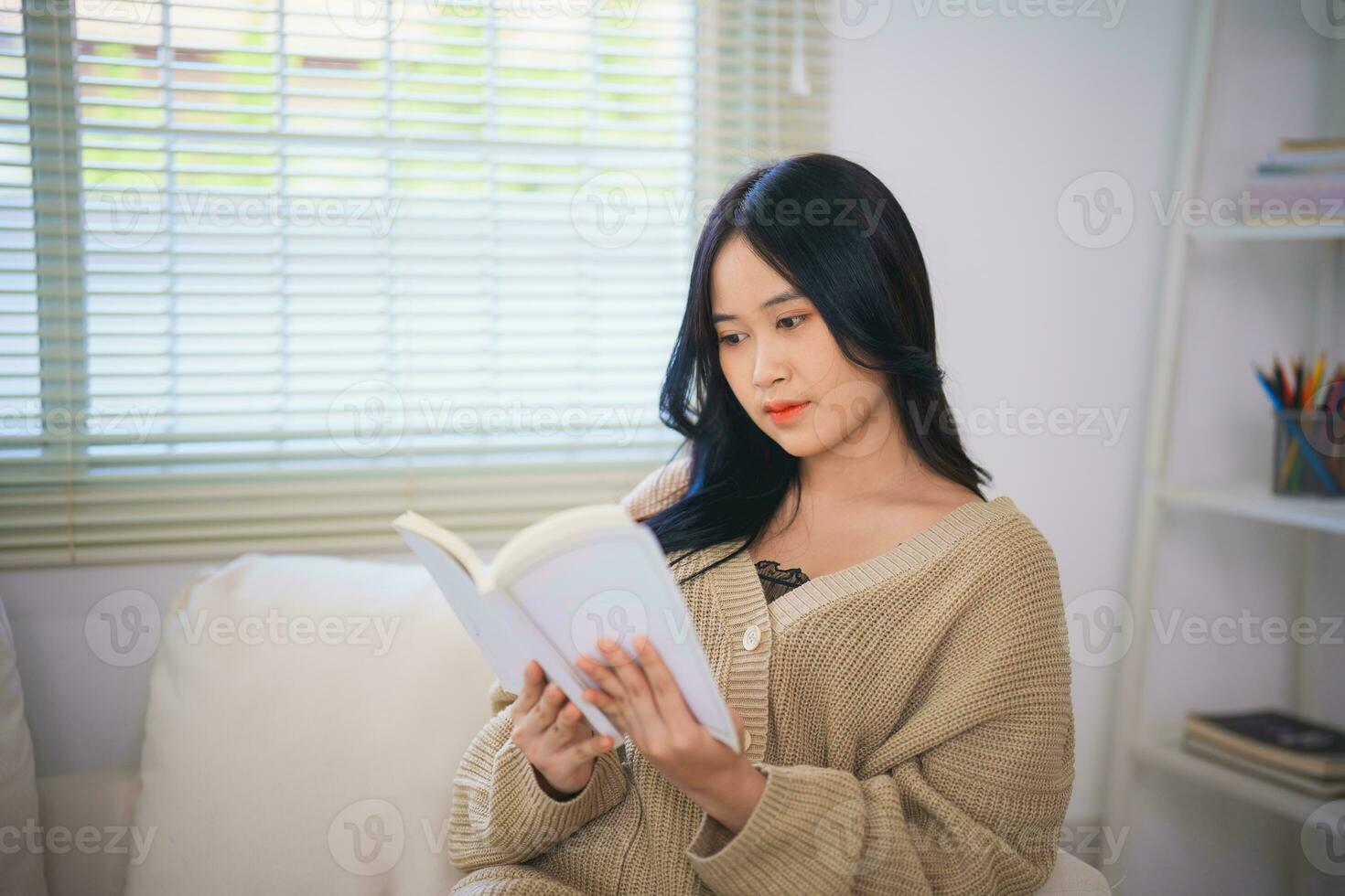 glad Lycklig asiatisk kvinna leende och läsning bok medan Sammanträde på soffa soffa i levande rum på Hem. asiatisk kvinnor koppla av läsning bok leende på soffa i de hus. aktivitet hobby på hus begrepp. foto